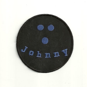 Pode incluir: Etiqueta redonda preta com olhos e boca bordados em azul, e o nome "Johnny" escrito abaixo.