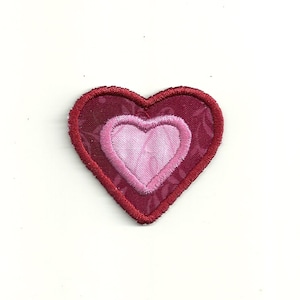 May include: A red heart-shaped patch with a smaller pink heart inside. The patch is made of fabric and has a stitched edge.