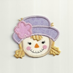 May include: A purple and pink felt applique of a scarecrow's face with a flower on the hat.