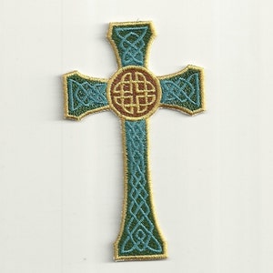 Peut inclure: Écusson brodé en forme de croix celtique, avec des détails turquoise et dorés. La croix présente un motif circulaire central marron et doré, avec des motifs de nœuds complexes. Les bords sont soulignés en or.