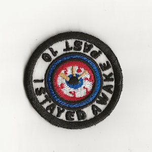 May include: A black and white embroidered patch with a clock face in the center. The clock face has red, white, and blue rings. The text around the clock face reads "Stay Up Past 10 To Awake".
