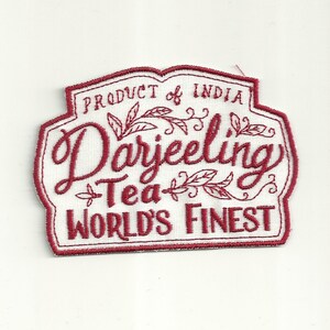 Puede incluir: Parche bordado rojo sobre fondo de tela blanca. El parche presenta el texto "Product of India Darjeeling Tea World's Finest" con diseños decorativos de hojas.
