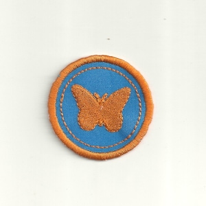 May include: Orange butterfly embroidered on a blue fabric circle with an orange stitched border.