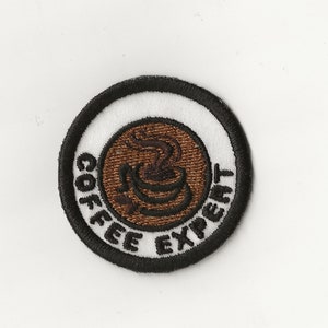 Könnte beinhalten: Ein weißer und schwarzer gestickter Aufnäher mit einer braunen Kaffeetasse und Dampf. Der Text "COFFEE EXPERT" ist um die Tasse herum geschrieben.