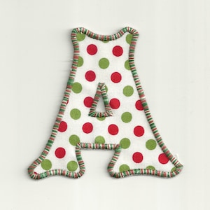 May include: A felt letter A with a white background and red and green polka dots. The letter is outlined in green and brown thread.