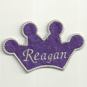 Pode incluir: Coroa de feltro roxo com acabamento de glitter prateado e o nome "Reagan" bordado em fio branco.