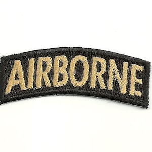Puede incluir: Parche bordado con la palabra "AIRBORNE" en letras doradas sobre un fondo negro curvado. El parche tiene un borde negro y está diseñado para ser cosido en ropa o equipo.