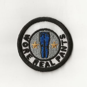May include: A round, black and white embroidered patch with the text "Work Real Pants" and a pair of blue jeans with two gold stars.