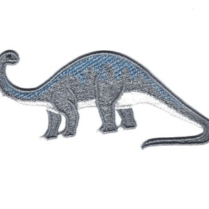 Può includere: Un toppa ricamata di dinosauro grigia e blu. Il dinosauro è in piedi su tutte e quattro le zampe con la coda arrotolata dietro di lui.
