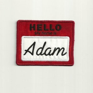 May include: Red felt name tag with white border and white background. The text "HELLO MY NAME IS" is printed in black on the top of the tag. The name "Adam" is written in black cursive on the white background.