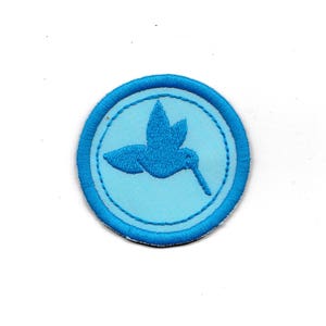 Könnte beinhalten: Ein blauer gestickter Aufnäher mit hellblauem Hintergrund, der einen Kolibri im Flug zeigt. Der Aufnäher ist rund mit einem blauen Rand.