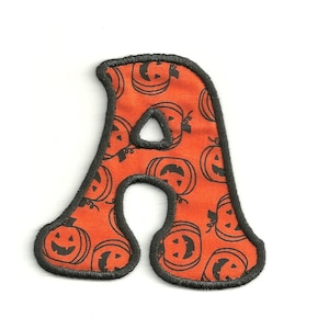 Peut inclure: Une lettre A en feutre avec un contour noir. La lettre est orange et présente un motif de visages de citrouilles noires.