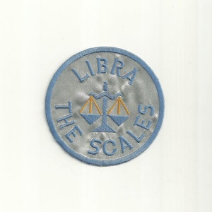 以下が含まれることがあります： 「LIBRA THE SCALES」という文字と、スタイリッシュな天秤の画像が刺繍された青と銀のワッペンです。