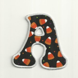 Peut inclure: Une lettre A en tissu noir avec un contour blanc. Le tissu est à motifs de maïs sucré orange et blanc sur fond noir.