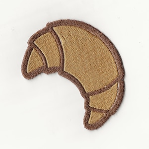 May include: Embroidered patch in the shape of a croissant. The patch features a light brown interior with a darker brown outline and details. The patch is on a white background.
