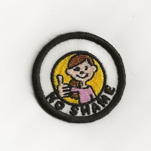 Pode incluir: Um remendo bordado branco e preto com fundo amarelo. O remendo apresenta uma menina de desenho animado com cabelo castanho dando um polegar para cima. O texto "No Shame" está escrito ao redor da borda do remendo.