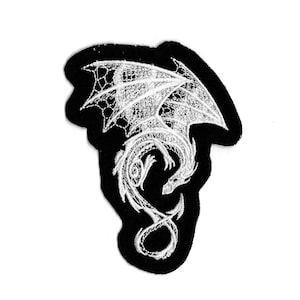 Może przedstawiać: Czarna łatka z haftowanym wzorem białego smoka. Smok jest zwinięty z rozłożonymi skrzydłami.