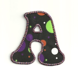 Peut inclure: Une lettre A en tissu noir avec un motif de pois de couleur violette, verte, orange et blanche. La lettre est bordée d'une garniture violette, orange et noire.
