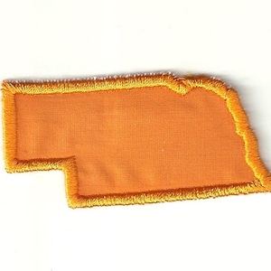 May include: An orange embroidered patch in the shape of the state of Nebraska. The patch has a gold border with a fringe effect. The background is a solid orange colour. The patch is on a white background.