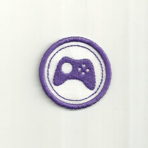 May include: A circular, embroidered patch featuring a purple video game controller icon within a white circle. The controller is centered, with a purple border and a white background. The patch is likely for clothing or accessories.