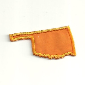May include: Orange felt outline of the state of Oklahoma with a yellow stitched border.