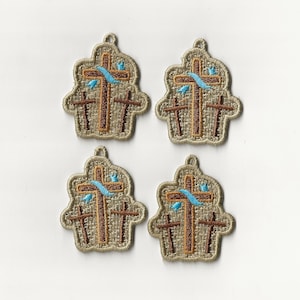 Op de afbeelding: Vier beige stoffen patches met geborduurde bruine kruisen en een blauw lint. De kruisen zijn verticaal gerangschikt met het bovenste kruis met een blauw lint eromheen.