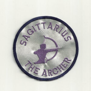 May include: A silver and purple embroidered patch with the words "Sagittarius The Archer" around the edge. The center of the patch features a purple silhouette of an archer with a bow and arrow.