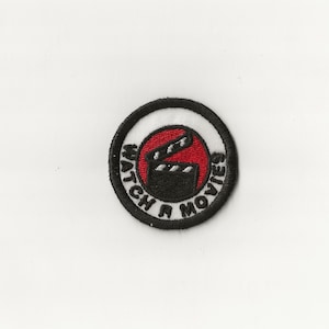 May include: A black and white embroidered patch with a red circle in the center. The circle contains a black and white film clapboard. The text "WATCH N MOVIES" is written in a circle around the clapboard.