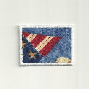 Puede incluir: Un pequeño parche cuadrado de tela con un borde blanco. El parche presenta un fondo azul con estrellas blancas y una bandera estadounidense roja, blanca y azul.