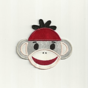 Könnte beinhalten: Ein graues Filz-Affen-Gesicht mit einem roten Hut und schwarzen Haaren. Der Affe lächelt mit einem roten Mund und weißen Zähnen.