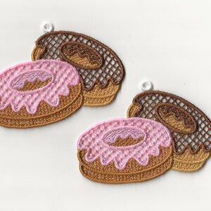 Op de afbeelding: Twee geborduurde donut-charms met roze en bruine glazuur. De donuts zijn bruin met een witte lus om op te hangen.
