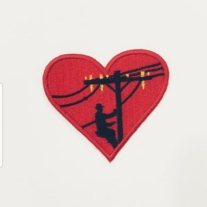 May include: Red heart-shaped embroidered patch featuring a silhouette of a person working on a power line. The power line has several yellow insulators.