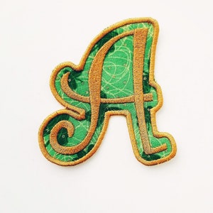 May include: A green and gold fabric patch in the shape of the letter A. The letter is outlined in gold thread and the fabric has a green and white pattern.