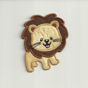 Puede incluir: Una aplicación de león bordada en marrón y beige con una cara sonriente. El león tiene una melena marrón y una lengua rosa.