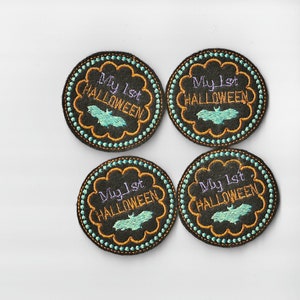 Può includere: Quattro cerchi in feltro nero con cuciture arancioni e turchesi. Ogni cerchio ha il testo "My 1st HALLOWEEN" in ricamo viola e arancione con un pipistrello turchese al centro.