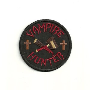 May include: A black embroidered patch with the words "Vampire Hunter" in red lettering. A crossed hammer and stake are in the center of the patch, with two brown crosses on either side.