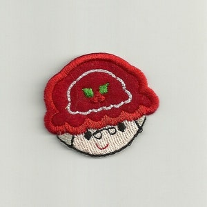 May include: Embroidered patch featuring a cartoon character with a red hat and a smiling face. The hat has a white trim and a small green and red berry design. The character's face is beige with black eyes and a black outline.
