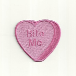 Może przedstawiać: Różowy łatkowy lizak w kształcie serca z haftowanym napisem "Bite Me" w kolorze białym.