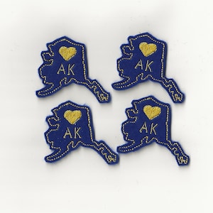 May include: Four blue felt patches shaped like the state of Alaska with gold embroidered outlines. Each patch has a gold heart with the letters "AK" inside and the words "I love AK" above the heart.