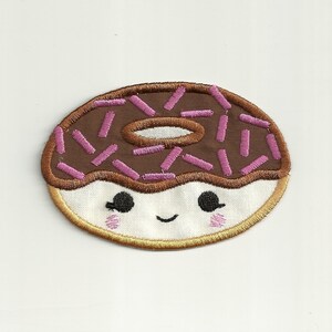 Op de afbeelding: Een bruine chocolade donut met roze sprinkles en een wit glazuur gezicht met lachende ogen.