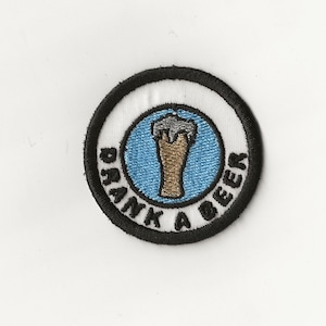 May include: A white embroidered patch with a black border. The patch features a blue circle with a white beer glass inside. The text "Drank A Beer" is written around the circle.