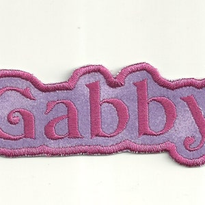 Könnte beinhalten: Lila und rosa gestickter Bügelaufkleber mit dem Namen "Gabby" in einer kursivschrift.
