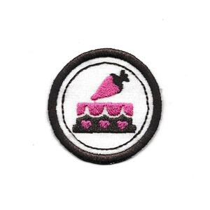 Peut inclure: Écusson brodé circulaire avec un motif de gâteau et de carotte. Le gâteau est rose et marron, avec des détails en forme de cœur. La carotte est rose. Le patch a un fond blanc et une bordure noire.