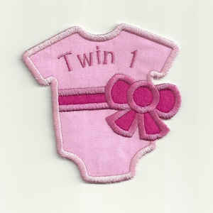 Puede incluir: Un body de bebé de tela rosa con un lazo rosa y el texto "Twin 1" bordado en la parte delantera.