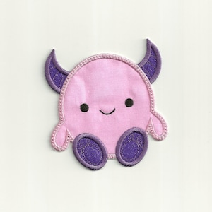Puede incluir: Un parche bordado de un monstruo lindo con cuernos y pies grandes, en rosa y morado.