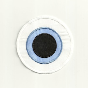 Könnte beinhalten: Ein weißer Stoffkreis mit einem blauen und schwarzen Augenmotiv. Das Auge ist von einem weißen Kreis mit einem blauen Ring und einer schwarzen Mitte umgeben.