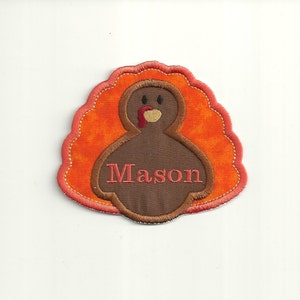 Op de afbeelding: Een bruine kalkoen met een rode keellap en een naamsticker met de tekst "Mason". De kalkoen wordt omringd door een oranje en rode gevederde staart.
