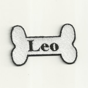 Könnte beinhalten: Weißer knochenförmiger Patch mit schwarzem gesticktem Text, der "Leo" lautet.