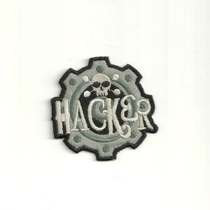 Peut inclure: Un écusson brodé gris et blanc avec un motif d'engrenage et le mot "HACKER" en lettres blanches. Un crâne avec des os croisés se trouve au centre de l'engrenage.