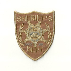 以下が含まれることがあります： ベージュの刺繍パッチで、金色の保安官の星と「SHERIFF'S SINCE 1878 DEPT.」の文字が刺繍されています